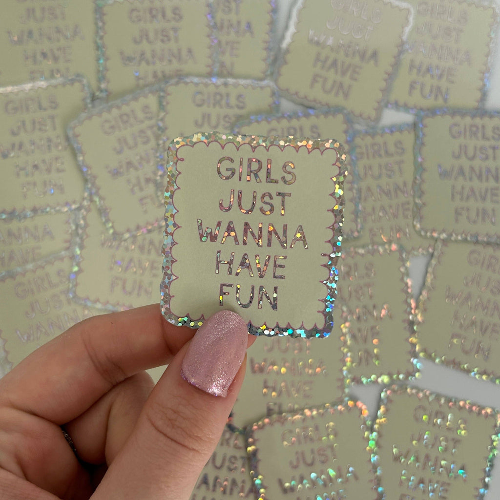 Holographic 'GIRLS JUST WANNA HAVE FUN' sticker held by a hand with pink nail polish against a background of similar stickers.