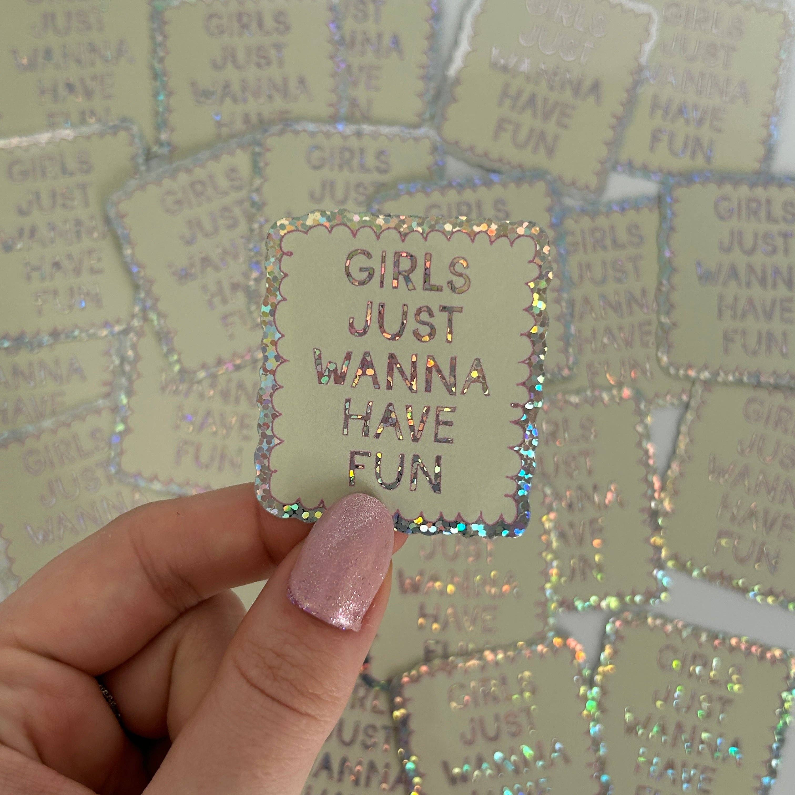 Holographic 'GIRLS JUST WANNA HAVE FUN' sticker held by a hand with pink nail polish against a background of similar stickers.